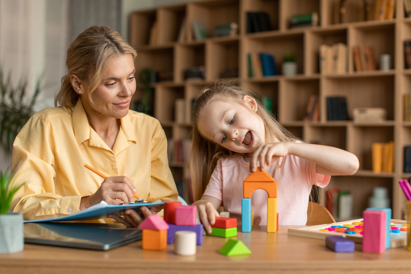 Child psychology exam. Little girl playing with colorful wooden blocks, professional kids psychologist writing notes